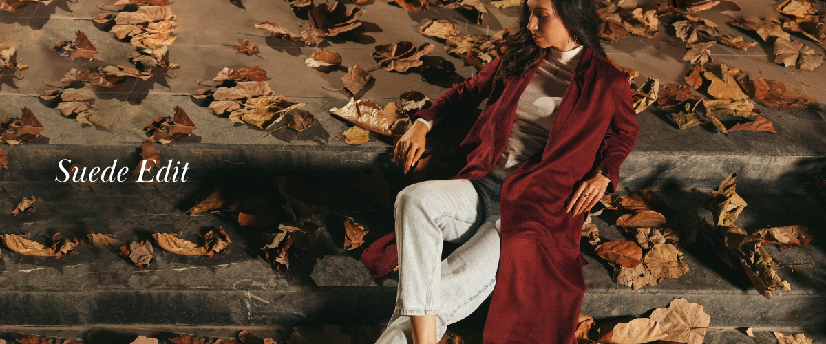 Woman wearing a long burgundy suede coat over a white turtleneck and light-wash denim, seated on stone steps covered in autumn leaves for the Cherie by Zeitgeist Suede Edit.