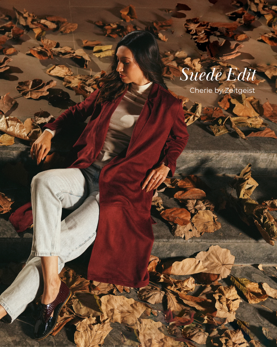 Woman wearing a long burgundy suede coat over a white turtleneck and light-wash denim, seated on stone steps covered in autumn leaves for the Cherie by Zeitgeist Suede Edit.